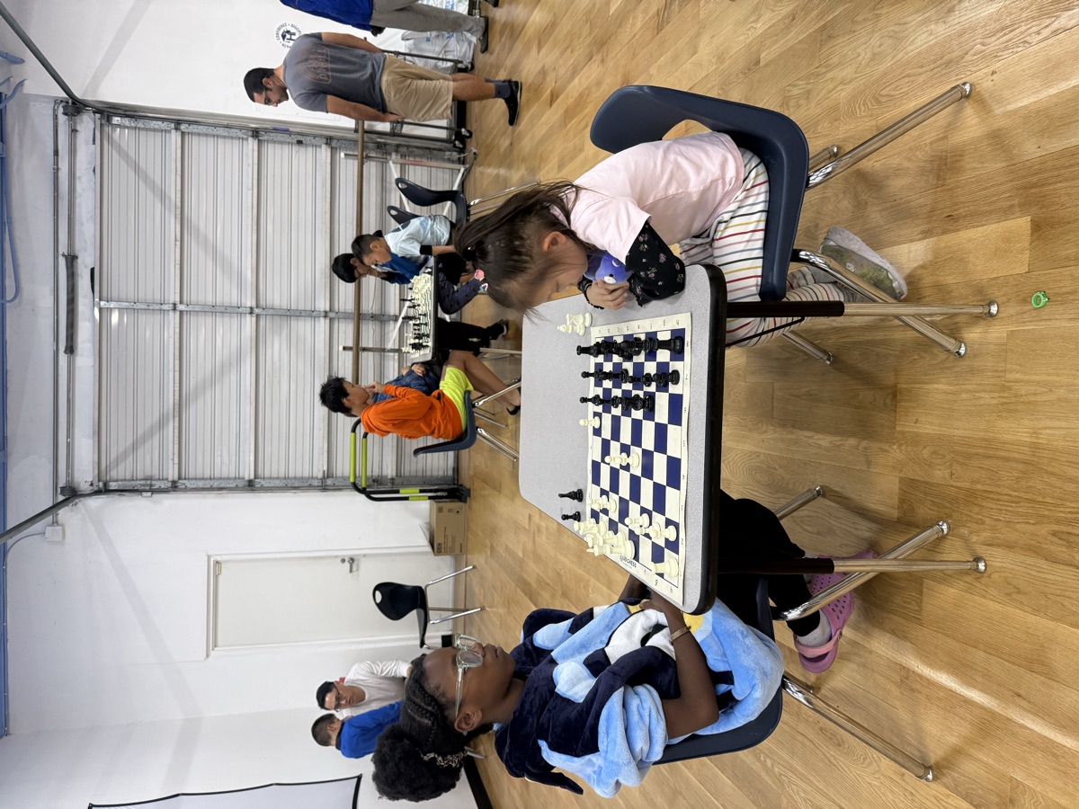 Two students facing off over a chess board while others play in the background
